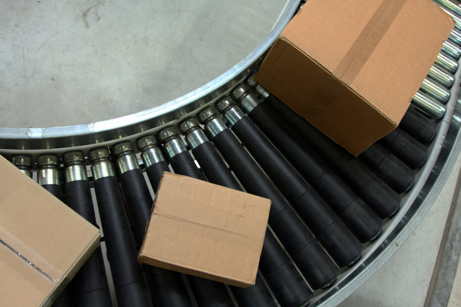 Three cardboard boxes placed on a curved section of a roller conveyor system with black rollers and metal frame, used for material handling in a warehouse setting.