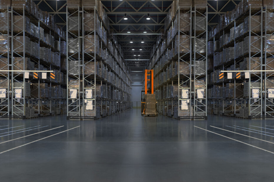 Wide view of a large warehouse with tall racks filled with palletized goods, organized in multiple rows. A yellow automated forklift or stacker is positioned in the center aisle, surrounded by clear, polished concrete flooring.