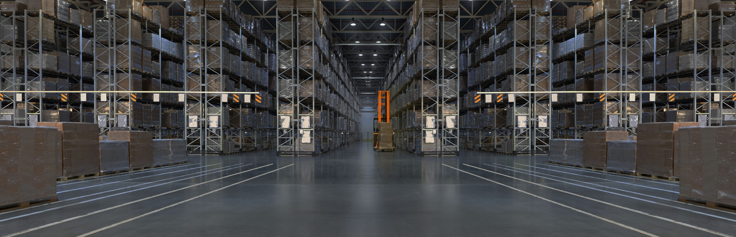 Wide view of a large warehouse with tall racks filled with palletized goods, organized in multiple rows. A yellow automated forklift or stacker is positioned in the center aisle, surrounded by clear, polished concrete flooring.