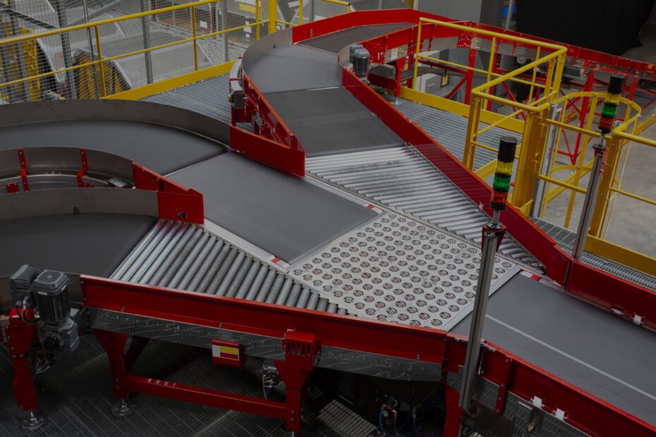 Conveyor sorting belt with red frames and black belts, featuring curved and straight sections, roller segments, and a transfer plate. Surrounded by yellow safety railings in a warehouse setting.