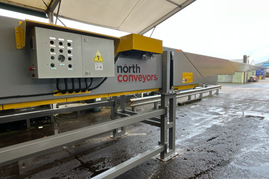 Outdoor view of a telescopic boom conveyor under a canopy, featuring a grey and yellow frame with an attached control panel displaying buttons and warning labels. The conveyor is branded with the text ‘north conveyors’ and is positioned on a wet concrete surface with industrial buildings visible in the background.