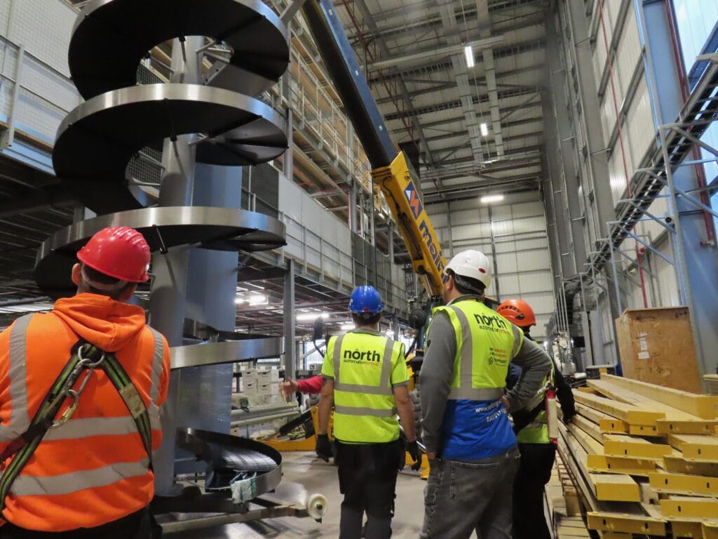 A group of men working together to install a spiral conveyor into a warehouse. The men are wearing hi-vis, some of which have the North Automation Group logo on the back. All of the men are wearing hard hats too.